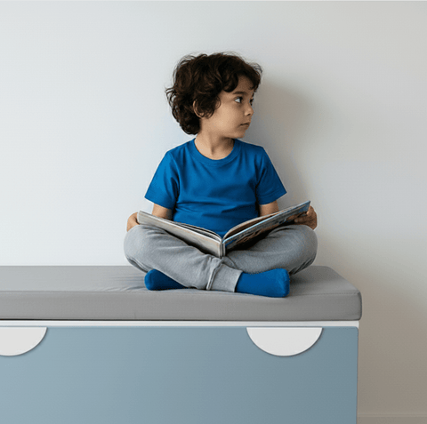 Ein kleines Kind mit lockigem Haar, das ein blaues Hemd und eine graue Hose trägt, sitzt im Schneidersitz auf einer Bank, hält ein aufgeschlagenes Buch in der Hand und blickt zur Seite gegen eine schlichte Wand.