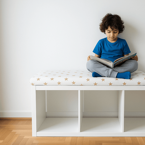Ein kleines Kind in einem blauen Hemd und einer grauen Hose sitzt im Schneidersitz auf einer weißen Bank mit goldenen Sternen und liest ein Buch. Unter der Bank befinden sich drei leere Regalfächer.