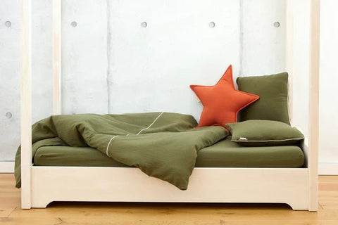 Ein minimalistisches Bett mit grüner Bettwäsche, einem passenden Kissen und einem orangefarbenen sternförmigen Kissen vor einer hellgrauen Wand und einem Holzboden.