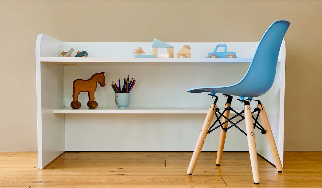 Ein kleiner weißer Schreibtisch mit Holzspielzeug, Buntstiften in einer Tasse und einem blauen Stuhl mit Holzbeinen auf einem Holzboden vor einer beigen Wand.