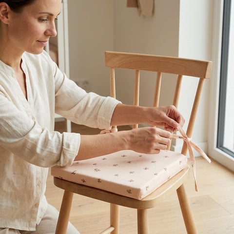 Eine Frau in heller Kleidung bindet ein Band, um ein rosa, gemustertes Sitzkissen auf einem Holzstuhl in der Nähe eines Fensters in einem hellen Raum zu befestigen.