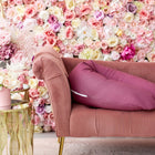 Ein rosafarbenes Samtsofa mit ergonomischem Design und einem KraftKids qualitäts Stillkissen Leinen magenta dunkel steht neben einem goldenen Beistelltisch mit rosa und weißen Blumen vor einer mit bunten Rosen geschmückten Wand.