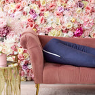 Eine rosafarbene Chaiselongue aus Samt steht vor einer bunten Blumenwand. Auf der Liege liegt ein KraftKids qualitäts Stillkissen Leinen in blau dunkelblau, und daneben steht ein goldener Beistelltisch mit pastellfarbenen Blumenarrangements in rosa Vasen.