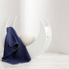 Eine halbmondförmige weiße Wiege mit einem runden cremefarbenen Kissen wird von einer KraftKids Babydecke Leinen blau dunkelblau geschmückt, die über eine Seite drapiert ist, vor weißen vertikal gestreiften Vorhängen und einem hellen Holzboden.