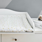 Auf einem weißen Wickeltisch liegt die KraftKids Wickelauflage Musselin gelbe Zitronen, deren gepolsterte Matte mit einem weißen Bezug mit kleinen gelben Zitronen versehen ist. Eine gefaltete Decke und weicher Schafsplüsch liegen auf der Kommode vor einer Wand mit geometrischem Muster.