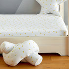 Ein weißes Kinderbett mit den KraftKids Spannbettlaken Musselin gelbe Zitronen mit einem kleinen gelben Zitronenmuster, einem passenden sternförmigen Kissen und einem weichen geknüpften Kissen auf einem Holzboden. An der Seite hängt ein heller Vorhang mit demselben Muster.