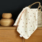 Eine cremefarbene KraftKids Babydecke Musselin Kirschen mit kleinen Kirschenaufdrucken drapiert sich über einen geflochtenen Korb neben zwei gestapelten Weidengefäßen auf einem Holzschrank vor einem schwarzen Hintergrund.