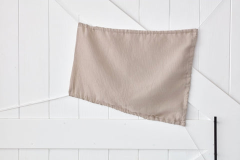 Der KraftKids Sonnensegel Musselin beige, leicht zerknittert und in einem sanften Beigeton, wird diagonal an eine weiße holzgetäfelte Wand mit sich kreuzenden Linien gepinnt.