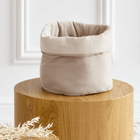 Das KraftKids Körbchen rund Musselin beige hat einen weichen, gefalteten Rand und steht stilvoll auf einem Holzsockel in einem minimalistischen weißen Raum. Getrocknetes Laub in der Nähe macht es ideal als Wickeltisch Organizer oder dekoratives Aufbewahrungskörbchen.