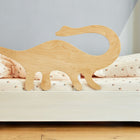 Ein Rausfallschutz aus Leimholz Dino von KraftKids steht auf einem cremefarbenen Bett mit geblümten Laken und Kissen neben einer hellgrauen Wand und einem Holzboden - perfekt für zusätzliche Sicherheit im Kinderzimmer.