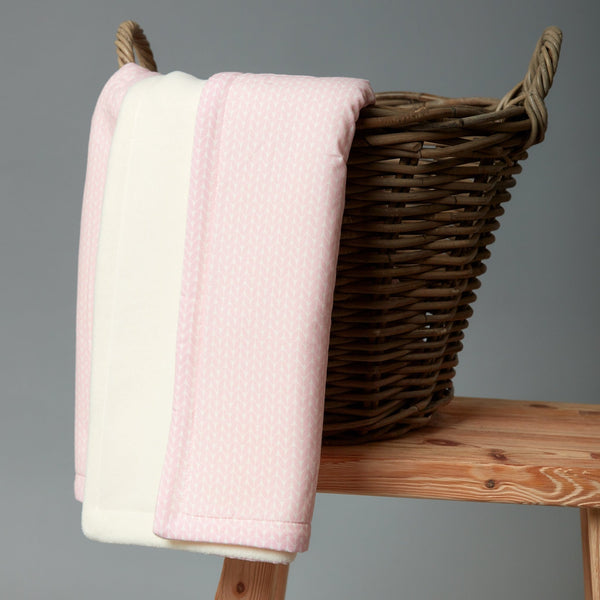 Ein Weidenkorb auf Holz hält eine gefaltete KraftKids Babydecke kleine Blätter rosa auf Weiß. Diese weiche Baumwolldecke mit rosafarbenem Blattmuster sorgt für Wärme vor dem neutralen grauen Hintergrund.
