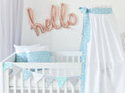 Ein weißes Babybett mit blau-weißer Bettwäsche aus 100 % Baumwolle, einem handgefertigten Betthimmel "Weiße Pfeile auf Blau" von KraftKids und einem pastellfarbenen Wimpel-Banner. Oben prangen roségoldene "Hallo"-Ballons vor einer weißen Wand - ideal für ein Himmelbett-Kinderzimmer.