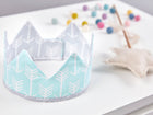 Eine handgenähte Stoffkrone von KraftKids mit weißem Pfeilmuster auf Mint liegt auf einer weißen Fläche. Im Hintergrund sorgen ein Stern-Zauberstab und eine bunte Filzkugel-Girlande für die perfekte Kinderparty-Stimmung.
