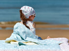 Ein junges Mädchen mit Kopftuch und Sonnenbrille sitzt auf der KraftKids Babydecke weiße Diamante auf Pastell Blau am Strand und blickt mit dem Rücken zur Kamera aufs Meer. Der Sandstrand und das ruhige blaue Wasser sind im Hintergrund zu sehen.