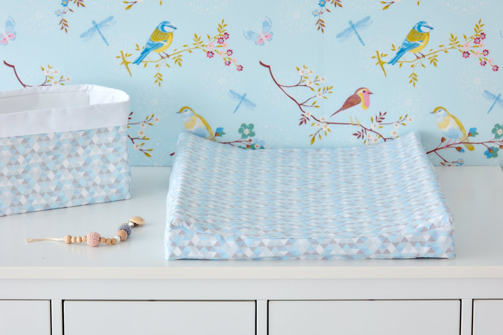 Ein KraftKids-Wickeltisch mit dem "Bezug für Keilwickelauflage kleine Dreiecke blau grau weiß", einer geometrischen blau-grau-weißen Wickelauflage, einem Aufbewahrungskorb und einer Schnullerklemme, vor einer Tapete mit bunten Vögeln und Libellen.