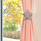 Eine Nahaufnahme einer rosafarbenen Gardine mit dem KraftKids "Gardinen Raffhalter weiße Punkte auf Grau", sternförmig mit weißen Punkten auf Grau, ist neben einem Fenster mit Blick auf Herbstbäume mit gelben und orangefarbenen Blättern zu sehen.