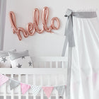 Ein weißes Babybett mit sternförmigen Kissen, einem Betthimmel Unigrau von KraftKids aus 100% Baumwolle und pastellfarbenen Wimpeln. Über der Krippe buchstabieren roségoldene Luftballons "Hallo" auf eine weiße Wand.