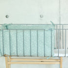 Ein minimalistisches Metall-Babybett mit KraftKids Bettumrandung Nestchen Musselin mint Pusteblumen, gefertigt aus 100% Baumwolle und mit einem zarten weißen Pusteblumen-Print, auf einem Holzständer vor einer schlichten Betonwand.