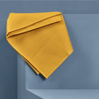 Die KraftKids Babydecke Waffel Piqué senf, eine strukturierte gelbe Baumwolldecke, ist ordentlich über den Rand einer geometrischen hellblauen Struktur vor einem passenden blauen Hintergrund drapiert.