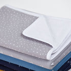 Ein ordentlich gefalteter Stapel Decken, wobei die oberste die Wickelunterlage Musselin grau Punkte von KraftKids ist - eine grau-weiß gepunktete Wickelunterlage mit einem weichen weißen Innenfutter, das teilweise sichtbar ist.