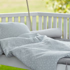 Ein gemütliches Outdoor-Bett mit dem KraftKids Bettwäscheset Musselin grau Pusteblumen aus 100% Baumwolle und Kissen sitzt auf einer weißen Holzschaukel und verspricht erholsamen Schlaf inmitten einer verschwommenen grünen Wiese und Bäumen.
