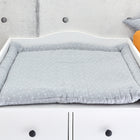 Die KraftKids Wickelauflage Musselin grau Pusteblumen ruht auf einer weißen Kommode an einer Betonwand, mit einer Aufbewahrungsbox, einem dekorativen Stern und einem kürbisförmigen Kissen im Hintergrund.