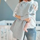 Ein Erwachsener hält ein in die KraftKids Babydecke Musselin grau Pusteblumen eingewickeltes Baby neben einem weißen Kinderbett in einem sanft beleuchteten Kinderzimmer. Nur die untere Hälfte des Gesichts des Erwachsenen und die Beine des Babys sind sichtbar.