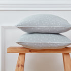 Zwei Kissenbezug Musselin grau Pusteblumen von KraftKids mit dezenten weißen Pusteblumenmustern sind auf einem Holzhocker vor einer weißen Wand mit Zierleisten gestapelt.