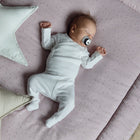 Ein Baby in Weiß schläft auf der weichen KraftKids Krabbeldecke Musselin goldene Punkte auf Rosa mit Polyestervlies-Füllung und hält einen Schnuller in der Hand. Daneben liegen zwei sternförmige KraftKids-Kissen, eines mintgrün und eines beige.