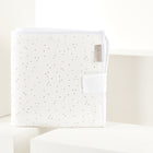 A KraftKids Reisewickelunterlage Musselin in Weiß mit goldenen Punkten steht auf weißen geometrischen Blöcken neben einem passenden Babybuch mit Stoffetikett, vor sanftem, neutralem Hintergrund.