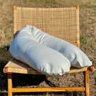 Ein großes, U-förmiges Stillkissen mit dem KraftKids Stillkissenbezug Leinen leicht grünes Weiß, passend Theraline 190 cm, wird auf einen geflochtenen Holzstuhl im Freien auf trockenem Gras gestellt.