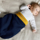 Ein Baby liegt in einem beigen Bettchen, trägt ein weißes langärmeliges Oberteil und den KraftKids Strampelsack Frühling Sommer Musselin dunkelblau und schaut mit erhobenem Arm zur Seite.