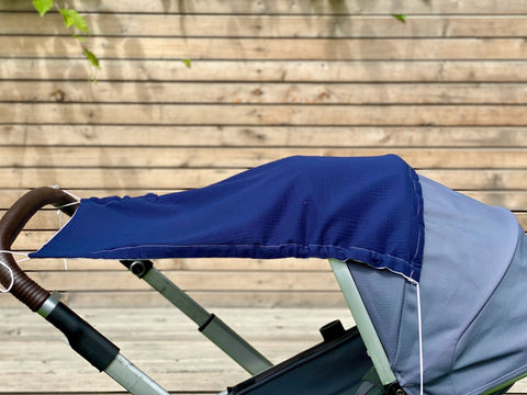 Nahaufnahme eines Kinderwagens mit dunkelblauem KraftKids Sonnensegel Musselin Verdeck, braunem Griff und hellgrauem Gestell vor Holzplanken und grünen Blättern. Das atmungsaktive Baumwollmaterial sorgt für Komfort und Schutz.