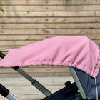 Ein KraftKids Sonnensegel Musselin rosa beschattet das Verdeck eines grauen Kinderwagens, mit einem Holzzaun und Grünpflanzen im Hintergrund.