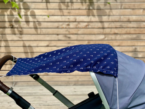 Ein KraftKids Sonnensegel Musselin dunkelblau Anker ist über ein Kinderwagenverdeck drapiert und zeigt seinen blauen, atmungsaktiven Musselin-Stoff mit weißem Ankerdruck. Im Hintergrund ist ein Holzzaun zu sehen, über dem blättrige Ranken wachsen.