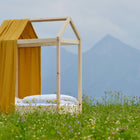 Ein einfaches Holzbettgestell mit dem KraftKids Bettbaldachin Musselin goldene Punkte auf Gelb steht in einer Wildblumenwiese, im Hintergrund sind Berge unter einem bewölkten Himmel zu sehen.