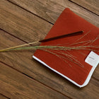 Eine KraftKids Reisewickelunterlage Doppelkrepp Rot Herbstrot mit weißem Rand und Etikett ruht auf Holz, begleitet von einem braunen Stift und getrocknetem Gras - ideal als platzsparende Wickelauflage für unterwegs.