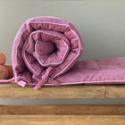 Eine aufgerollte KraftKids Bettumrandung Nestchen Cord Breitcord Rosa aus 100% Baumwolle mit Stoffbändern liegt auf einer Holzbank neben drei runden Holzbällen vor grauem Hintergrund.