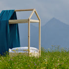 Eine KraftKids Bettbaldachin Musselin mit goldenen Punkten auf Petrol schmückt ein minimalistisches Holz-Bett im Wildblumenfeld vor Bergen - perfekte Inspiration für traumhafte Kinderzimmer-Deko.