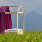 Ein KraftKids Bettbaldachin Musselin purpur ziert ein hölzernes Hausbett mit weißer Bettwäsche, eingebettet in ein Wildblumenfeld vor Bergen und Wolken - perfekt verträumte Kinderzimmer Deko.