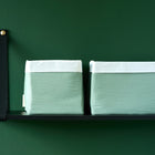 Zwei Körbchen Doppelkrepp Grün Jade von KraftKids mit weißen Aufsätzen sitzen auf einem schwarzen Wandregal vor einer dunkelgrünen Wand - stilvolle Aufbewahrung am Wickeltisch. Das Regal hat eine schwarze Metallhalterung und eine goldene Schraube.